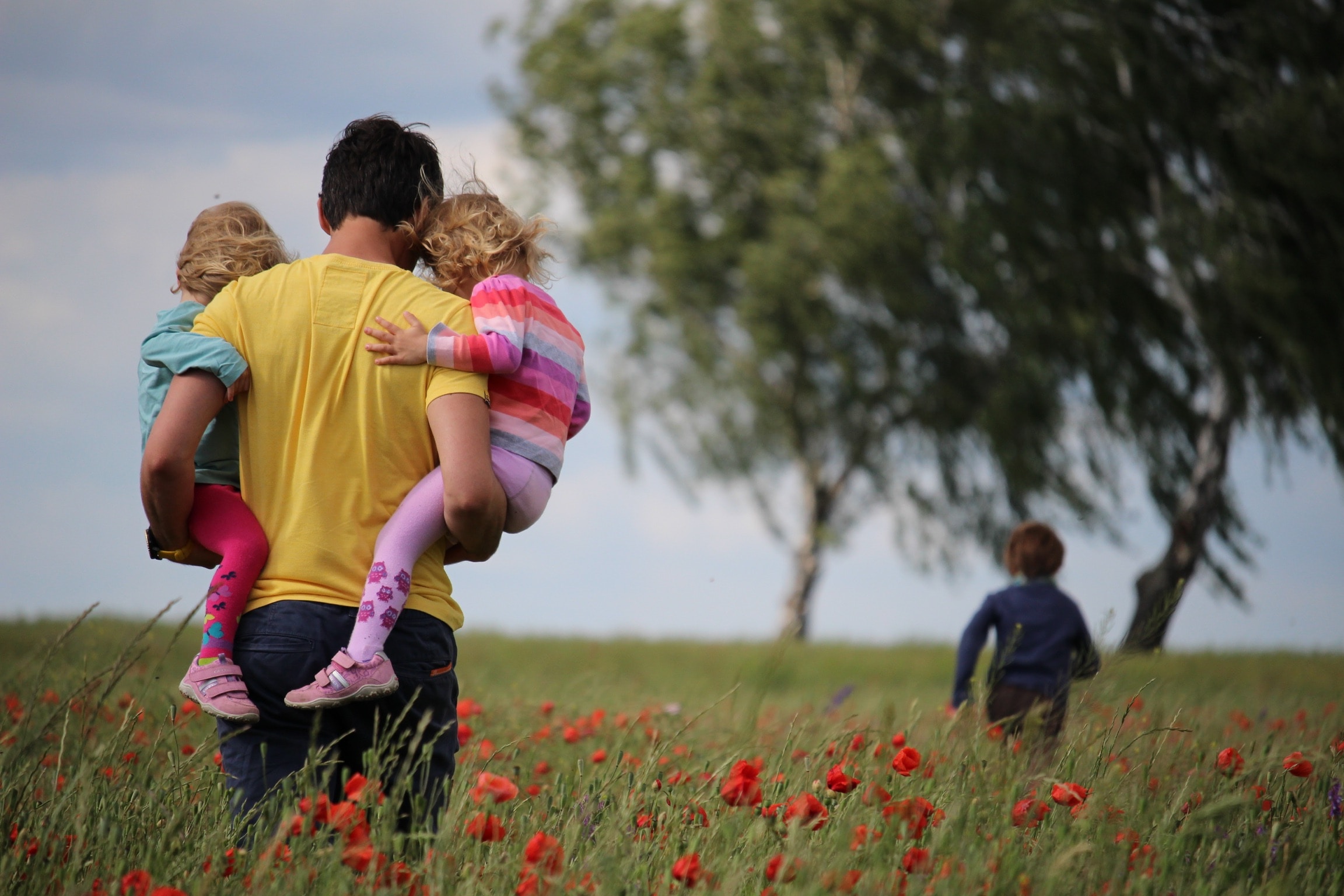 A parent carrying two children on their hips through a field with a tree. Credit: Photo by Juliane Liebermann on Unsplash